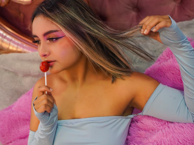 CloeesSuarez with pink eyeliner, blue off-shoulder top, and blonde hair, holding a red candy to her mouth, lying on a pink fluffy pillow, touching her hair.