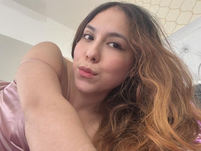 AmberlStha has light brown skin, wavy brown hair with blonde tips, and is pouting her pink lips. She's wearing a pink satin dress, showing her bare shoulder.