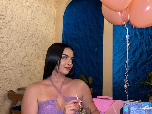 NovaDixon with long black hair, purple halter top, holding a drink, smiling, pink balloons, textured wall, and a pink bag on a table. Boobs visible, cute, sexy.