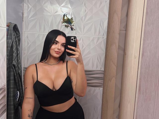 NovaDixon with black bra showing her big tits, holding a phone in a mirror selfie, long black hair, tattoo on right arm, black pants, necklace, smiling, bathroom.