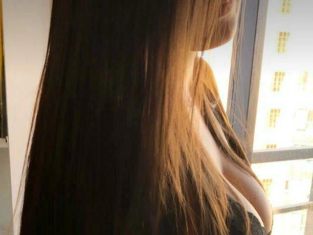 MicaelaMadrid-hot's long brown hair, slightly parted to reveal her bare shoulder and upper chest, stands in front of a sunlit window. Her back is to the camera.