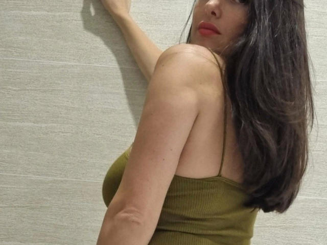MicaelaMadrid-hot with long brown hair, olive green tank top, red lipstick, and arm against textured wall, showing her tits and looking over shoulder.