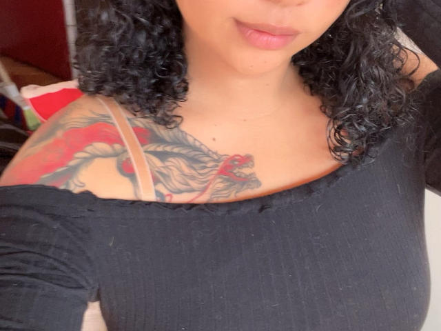OctaviaSteel with curly black hair, black off-shoulder top, revealing a colorful dragon tattoo on her right shoulder and upper chest, pink lips slightly puckered. Boobs partially visible.