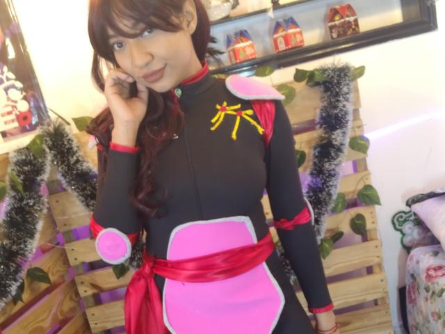 OctaviaSteel has dark brown hair, wearing a black and pink costume with red accents, yellow star, and pink shoulder pads, standing in a room with Christmas decorations.