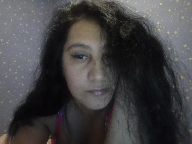 KarinaCecillia with long, messy black hair covering one eye, wearing a pink top, has light brown skin and subtle makeup, looking slightly to the side with a neutral expression.