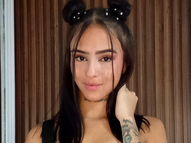 AlexiaBennet has black hair in double buns with beads, dark eyebrows, long lashes, pink lips, a tattooed arm, and a black choker, looking cute with a slight smile.