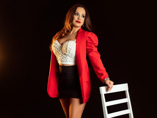 DashingFoxyX with big tits, wearing a red blazer, white patterned bustier, black skirt, standing, holding a white chair, dark background, red lipstick, long brown hair, confident smile.