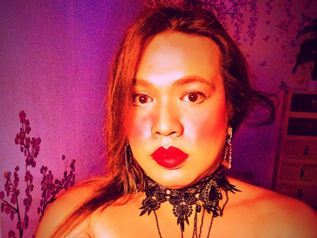 JadeWithLove has red lipstick, black lace choker, and earrings, with orange and purple lighting on her face, and her tits are partially visible. Her brown hair is loose.