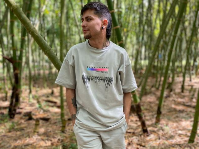 Sthefano with short black hair, earbuds in, wearing a white t-shirt and pants, standing in a bamboo forest, hands in pockets, looking to the side.