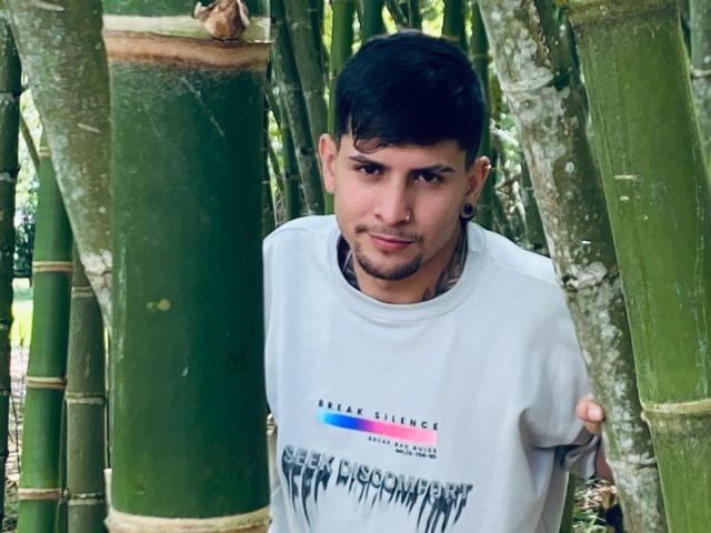 Sthefano with short black hair, small nose ring, and slight beard, wearing a gray shirt, holding green bamboo poles. Sthefano's face shows a subtle, confident smile.