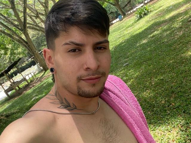 Sthefano with short dark hair, brown eyes, and a nose ring, has a tattoo on his neck, black ear gauges, and a pink towel over his shoulder, shirtless.