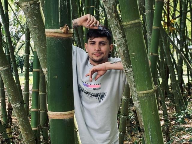 Sthefano with dark hair, light beard, and tattoos, leaning between green bamboo stalks, wearing a gray shirt, pointing with his right hand, smiling slightly.