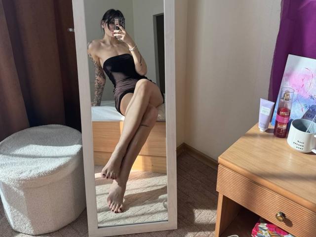 Tsukerberg con dei tatuaggi, che indossa un miniabito nero senza spalline, seduta su un letto di fronte a uno specchio, mostrando i piedi nudi e le unghie dei piedi dipinte di rosso, mentre tiene in mano un telefono..