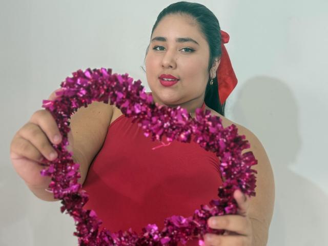ZoeGiil with dark hair, red lipstick, and red dress, holding pink heart with purple tinsel, looking at camera, red bow in hair, light skin, earrings.
