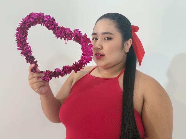 ZoeGiil with long black hair in a red halter top, holding a pink heart-shaped tinsel, wearing a red ribbon in her hair, smiling with red lipstick.