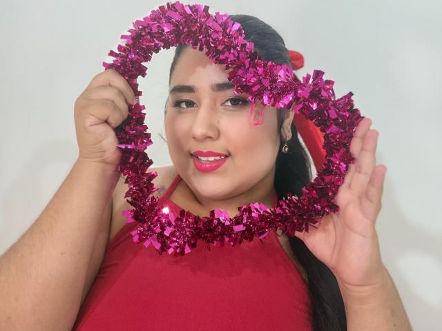 ZoeGiil holding a pink glittery wreath with both hands, wearing a red sleeveless top, smiling with pink lipstick, and dark hair in a red hairclip.
