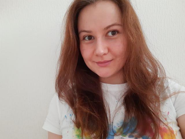 EvaNoNude tem cabelos castanhos ondulados e um tom de pele claro. Ela está vestindo uma camisa branca com manchas de água coloridas, sorrindo suavemente com seus olhos castanhos..