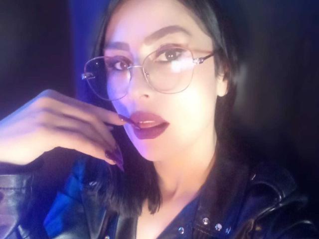 VioletaWhitee with glasses, dark lipstick, and black nails, wearing a leather jacket, touching her chin, has pale skin and straight black hair, looking at the camera.