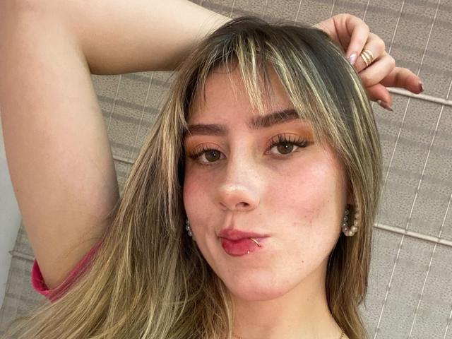 LivelyVanderWood tiene labios carnosos, cabello rubio con flequillo y un anillo en su mano derecha. Está sacando un poco la lengua, con una expresión linda y juguetona..