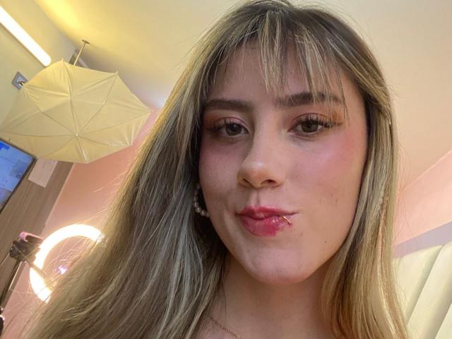 LivelyVanderWood tiene labios rosados y voluminosos, cabello rubio largo con flequillo, y está haciendo una mueca, con delineador de ojos rosa y maquillaje sutil, en una habitación bien iluminada..