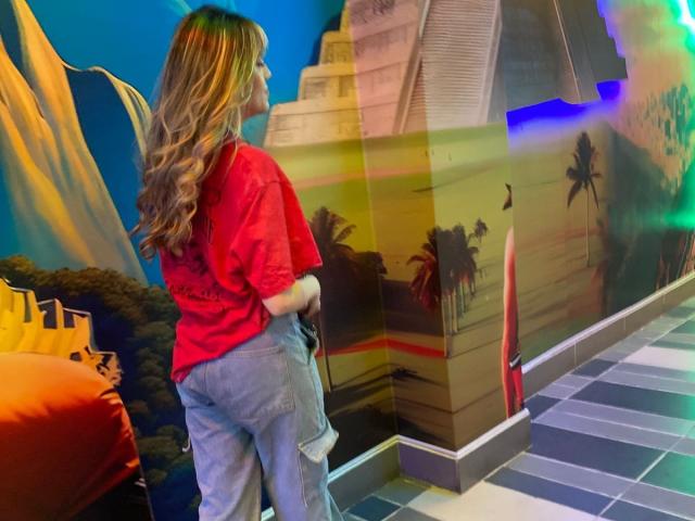 LivelyVanderWood está de espaldas a la cámara, vestida con una camisa roja y pantalones vaqueros azules, mostrando sus glúteos. Tiene el pelo ondulado, de color rubio a castaño..