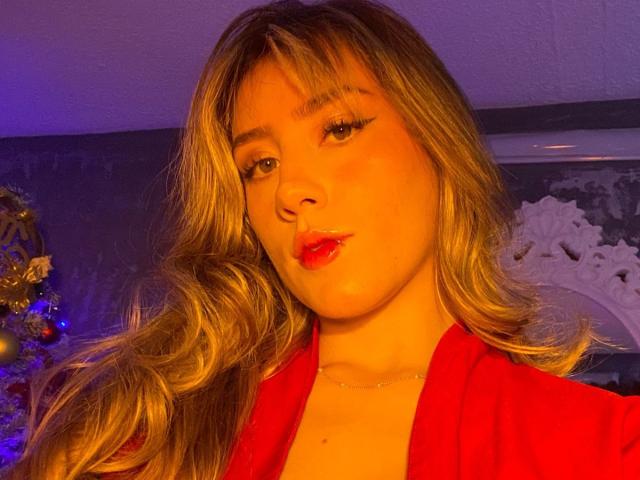 LivelyVanderWood, com cabelo loiro ondulado, batom vermelho e robe vermelho, olhando para a câmera com um leve franzido, em iluminação quente, amarelada-alaranjada. Seios não visíveis, foco no rosto..