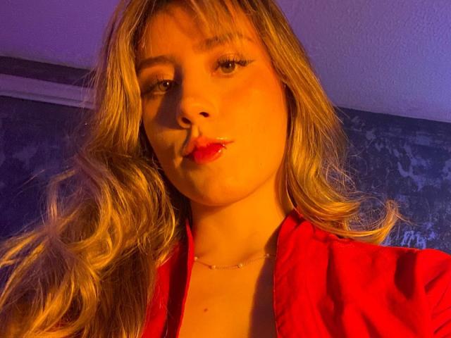 LivelyVanderWood, con cabello rubio ondulado, lápiz labial rojo y una camisa roja, haciendo pucheros con una ligera sonrisa, iluminada por una cálida luz amarilla y púrpura..