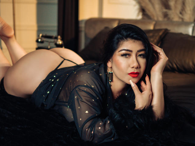 KhloeLawson is lying on a black fur blanket, wearing a sheer black robe with gold studs, red lipstick, and black thong, showing her ass, with her hand on her face.