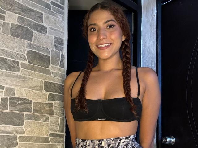 MiaBianchi with braided red hair, smiling, wearing a black sports bra, showing small tits, standing in a doorway, wearing high-waisted patterned pants.