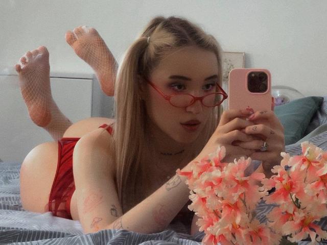 MillyPopa with red glasses, red lingerie, fishnet socks, lying on bed, taking selfie, showing ass, blonde pigtails, tattoos, pink phone case, pink flowers, ring on finger.