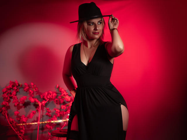 PerlaDivaio with blonde hair, black dress, and hat, showing a side slit, holding hat, red lighting, heart decoration, and flowers, standing confidently, showcasing her ass.