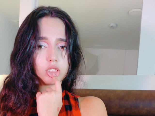 MelissaSmithers with wavy black hair, light pink eyeshadow, and a red plaid top, biting her finger, showing slightly parted pink lips, looking to the side in a selfie.