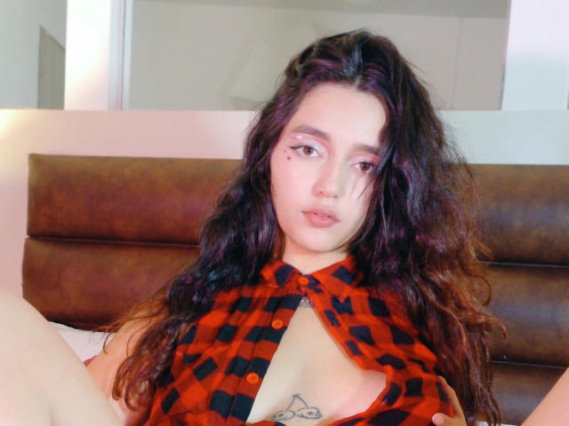MelissaSmithers with long, wavy dark hair, wearing an open red plaid shirt, exposing her tits and a small tattoo below her chest, sitting with legs spread.