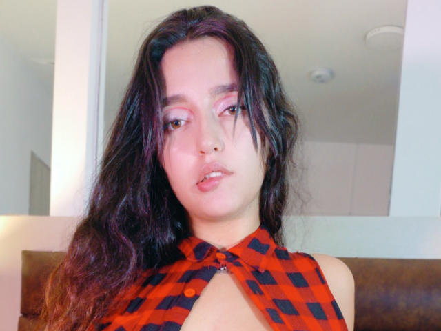 MelissaSmithers with long, wavy black hair, pink eyeshadow, and a red-black checkered halter top showing a small cutout, looking at the camera with parted lips.