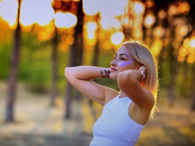 Lilitraya with blonde hair, purple face paint, white tank top, arms raised, tattoo on forearm, standing in sunlit forest, glowing golden light, peaceful expression, bokeh effect.
