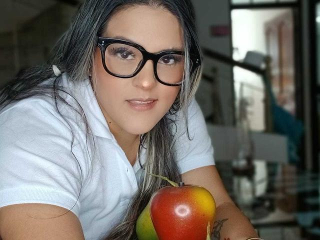 MiaJhonss con occhiali neri, capelli grigi lunghi, camicia bianca, che tiene una mela rosso-gialla, con un leggero sorriso, un tatuaggio sul braccio destro, che guarda la fotocamera, in un ambiente interno informale..