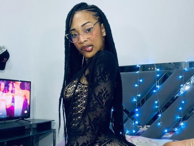 ElaHott with long black braids, wearing glasses and a black lace dress, sitting on a bed, showing her small tits and cute face, with blue string lights.