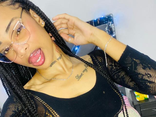 ElaHott has dark braids, glass frames, and a tongue out with saliva. She's wearing a black lace top, showing her tits and a tattoo under her collarbone.