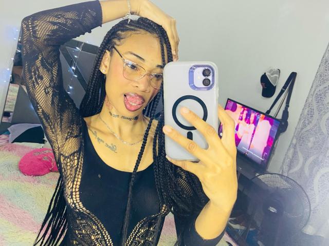 ElaHott with long braids, glasses, black lace top, tongue out, holding phone, black tank, tattoos, and pink bed in background, taking a mirror selfie.