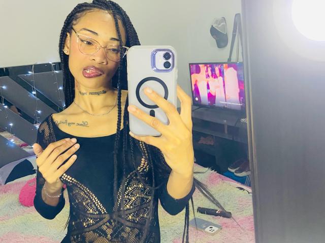 ElaHott with braided hair, glasses, and pink lipstick, wearing a black lace top, taking a mirror selfie with her phone, showing her small tits and choker.