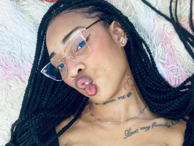 ElaHott with black braids, pink lipstick, puffed cheeks, and tongue out, wearing glasses, has tattoos on neck and chest, and a chain necklace, laying on white fluffy pillow.