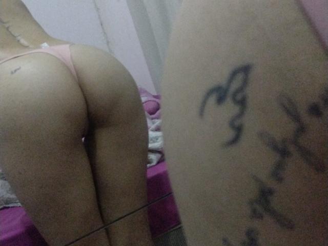 BrazilianWild is kneeling with her ass in pink panties, reflected in a mirror. Her tattooed arm is in the foreground. Her butt is prominently displayed.