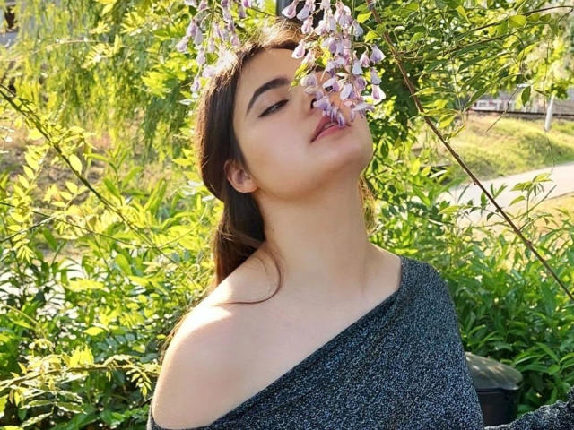 DeniseLove – sinnlich, mit leicht geneigtem Kopf, die violetten Blumen beschnuppernd, trägt sie einen grauen Pullover mit einem Schulterteil, ihr braunes Haar ist zu einem lockeren Pferdeschwanz gebunden, um sie herum grünes Laub..