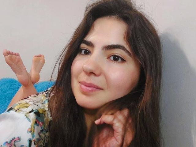 DeniseLove - aantrekkelijk, met lang bruin haar, bruine ogen en roze lippen, liggend, benen omhoog, gekleed in een bloemenprint shirt, zacht lachend, hand onder haar kin, blote voeten zichtbaar..