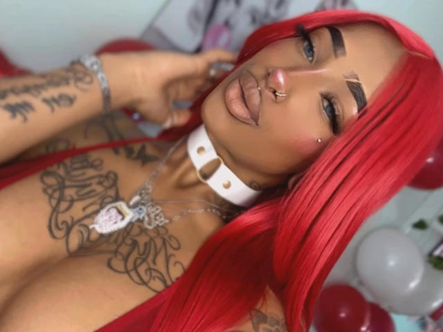 EvanYelin69 has bright red hair, tattoos, a white collar, nose ring, and a silver necklace, with her tongue out, showing her pouty lips and large tits.