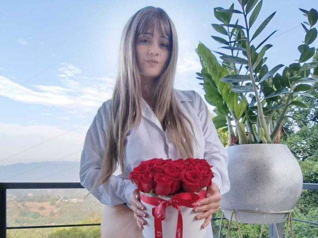 EmmaGoldyx mit langen blonden Haaren, die ein weißes Hemd trägt und einen Strauß roter Rosen hält, steht neben einer hohen Topfpflanze und lächelt sanft..