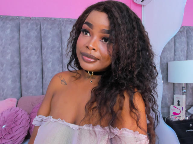 MadinsonLopez with curly black hair, brown skin, wearing an off-shoulder white top, black choker, and makeup, showing small tits, a tattoo on her left shoulder, and a cute smile.