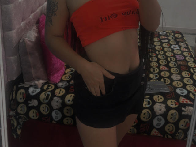 SarahGood in a red crop top, showing her flat stomach and black skirt with her hand in the pocket, has a tattoo on her arm and a pink pillow.