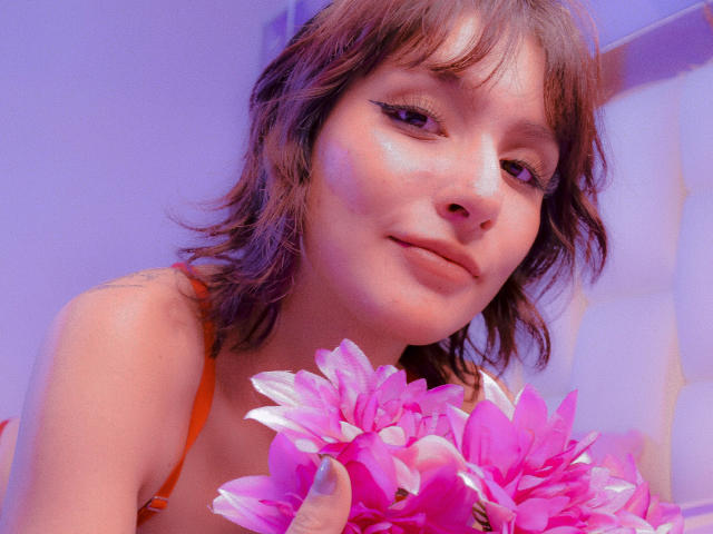 IsabellaHallad with wavy brown hair, subtle eyeliner, and pink flowers in her hand, smiling softly. She's wearing an orange top, and the image has a purple-blue tint.