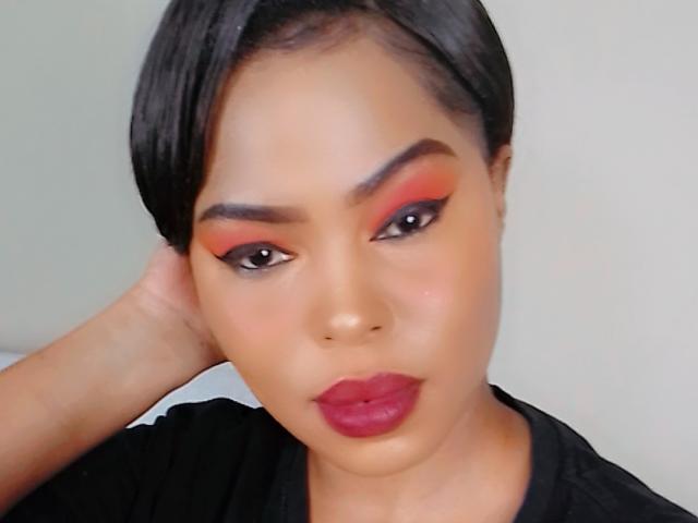 Sadeh69 with dark hair, red and orange eyeshadow, black eyebrows, pinkish skin, red lipstick, black top, hand behind head, looking at camera, slightly smiling.